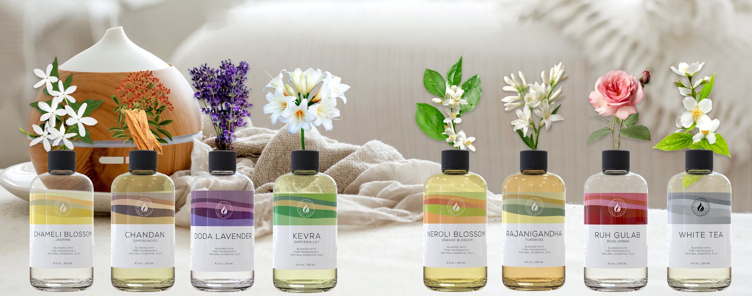 A full lineup of Aroma Country diffuser oils displayed with matching botanicals, including jasmine, sandalwood, lavender, gardenia, orange blossom, tuberose, rose, and white tea, representing each fragrance blend.