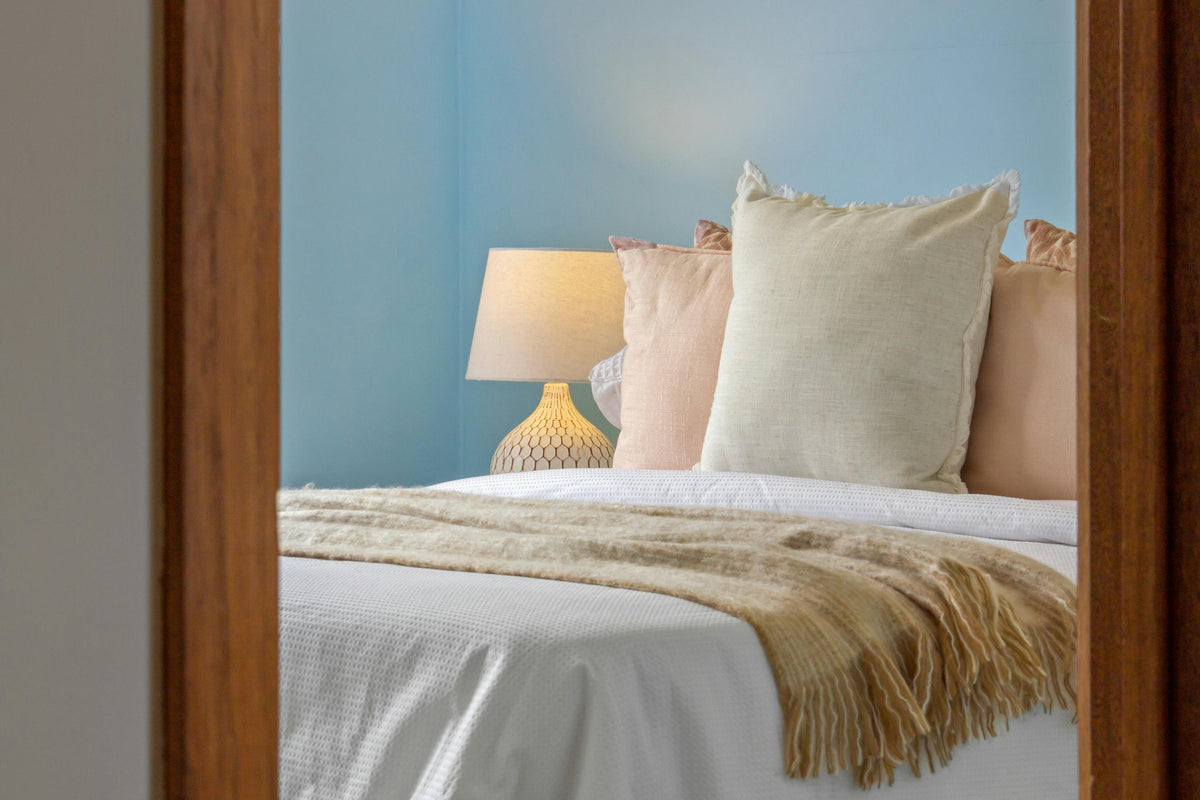 Inviting bedroom with layered neutral pillows, a textured beige throw, and a softly lit table lamp against a pale blue wall.