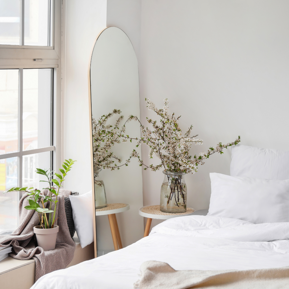 Serene bedroom with a white bed, large arched mirror, and vase of flowering branches on a round side table near the window.